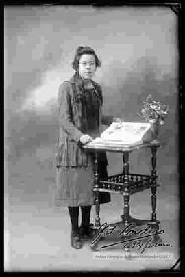 Foto de estudio de una dama de lentes que se encuentra parada y sosteniendo un libro que se encuentra en cima de una mesita. Lleva puesto un traje y vestido formal a rayas.