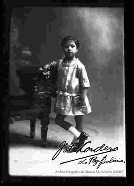 Foto de estudio de una niña apoyada a un sillón y sosteniendo en la mano derecha unas flores