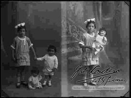 Foto de estudio de dos niñas que se encuentran tomadas de la mano y paradas lado a lado de una muñeca