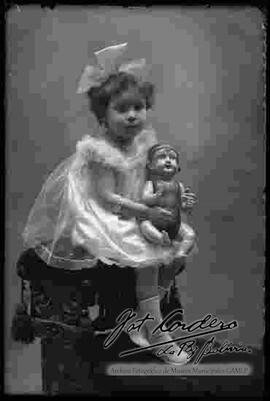 Foto de estudio de una niña que se encuentra sentada en la cabecera de un sillón. Se encuentra sujetando una muñeca