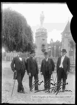 Cuatro varones vestidos de traje con sombreros bombím y delante del monumento a los Colorados de Bolivia al final de la alameda