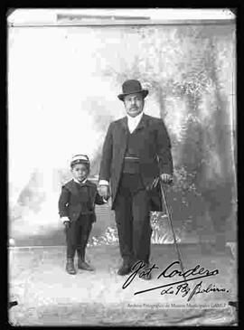 Foto de estudio de un varón que sostiene a su niño. Lleva puesto un sombrero bombín  y un traje con chaleco de color negro, sostiene en la mano izquierda unos guantes y un bastón