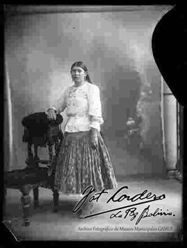 Foto de estudio de una chola que se encuentra parada y apoyada a un sillon. No lleva sombrero. Viste una blusa cerrada de manga larga, pollera floreada, botines, un collar con libra esterlina y aretes.