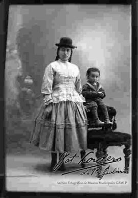 Foto de estudio de  una chola que lleva puesto sombrero borsalino de color oscuro, blusa con encajes y de manga larga, pollera y botines con tacón, parada a lado de un sillón donde se encuentra sentado su hijo pequeño