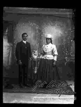 Foto de estudio de una pareja. Una chola que se encuentra parada junto a su esposo y apoyada a una mesita con flores. Lleva puesto un sombrero blanco de copa alta, una blusa blanca con encajes y moños, pollera y botines con tacón.