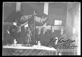 Grupo de 8 profesores y un cura, en el interior de un salón, detrás de una mesa y donde se puede apreciar de fondo la bandera de Bolivia