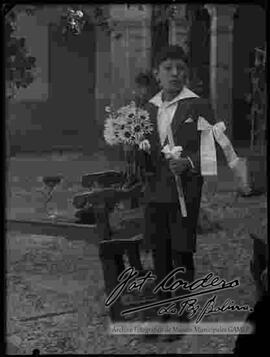 Niño en una casa particular, en el día de su primera comunión, sosteniendo una vela y un ramo de flores
