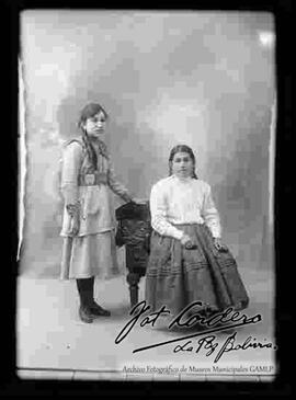 Foto de estudio de una chola que se encuentra sentada en un sillón, sin sombrero. Lleva puesto aretes, una blusa blanca de cuello cerrado y manga larga, pollera, botines con tacón y junto a su hija que se encuentra parada junto a ella.