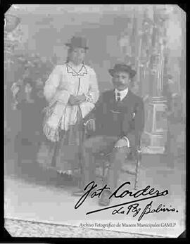 Foto de estudio de una pareja. Una chola que se encuentra parada y que luce un sombrero borsalino, blusa en cuello v, manta en brazo, pollera y zapatillas planas. El caballero es de bigotes con sombrero, traje y corbata, el mismo se halla sentado