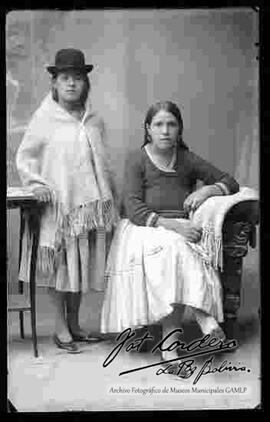 Foto de estudio de dos cholas. Una se encuentra parada y apoyada a una mesita, con sombrero borsalino, manta puesta, pollera y zapatillas. La otra se encuentra sentada en un sillón, viste una chompa, pollera, zapatillas planas y no lleva sombrero.