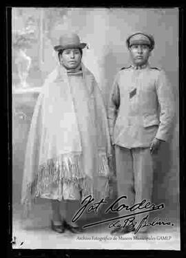 Foto de estudio de una pareja. Una chola que lleva puesto un sombrero borsalino, manta,  pollera y zapatillas planas. El varón presenta vestimenta de policía