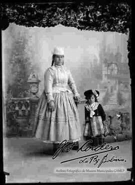 Foto de estudio de una chola que se encuentra parada y apoyada en la columna de una baranda, junto a su hija. Lleva puesto un sombrero blanco de copa alta, blusa de cuello cerrado, pollera con cetros sobresalidos y botines con tacón.