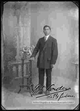 Foto de estudio de un joven que viste de traje y que se encuentra parado a lado de una mesita con flores