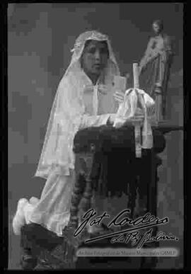 Foto de estudio de una niña el día de su primera comunión, de rodillas encima de un sillón, delante de la imagen del Corazón de Jesús
